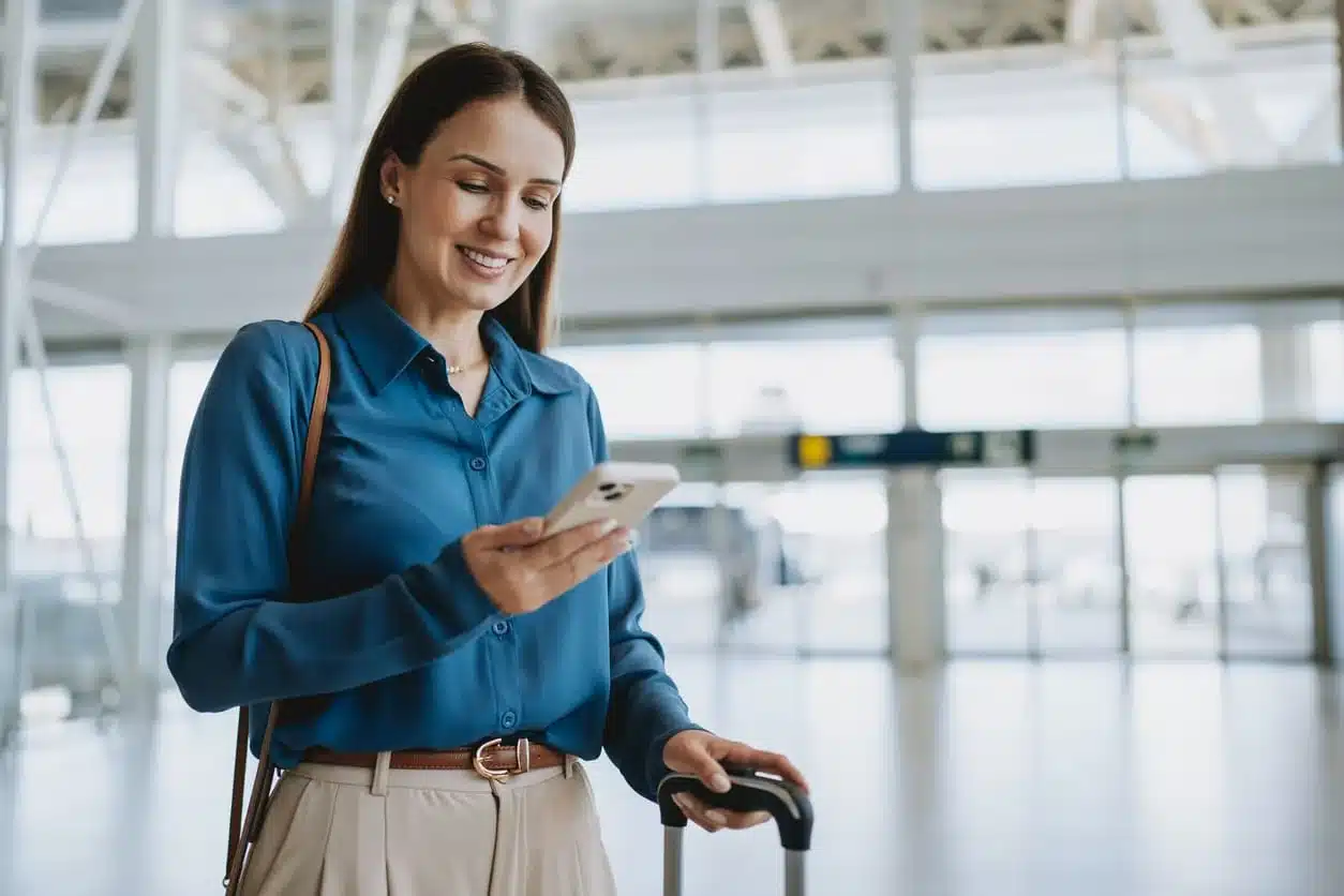 Woman in a blue shirt smiling at her phone in an airport terminal, standing beside a rolling suitcase as a permanent resident preparing to travel abroad.