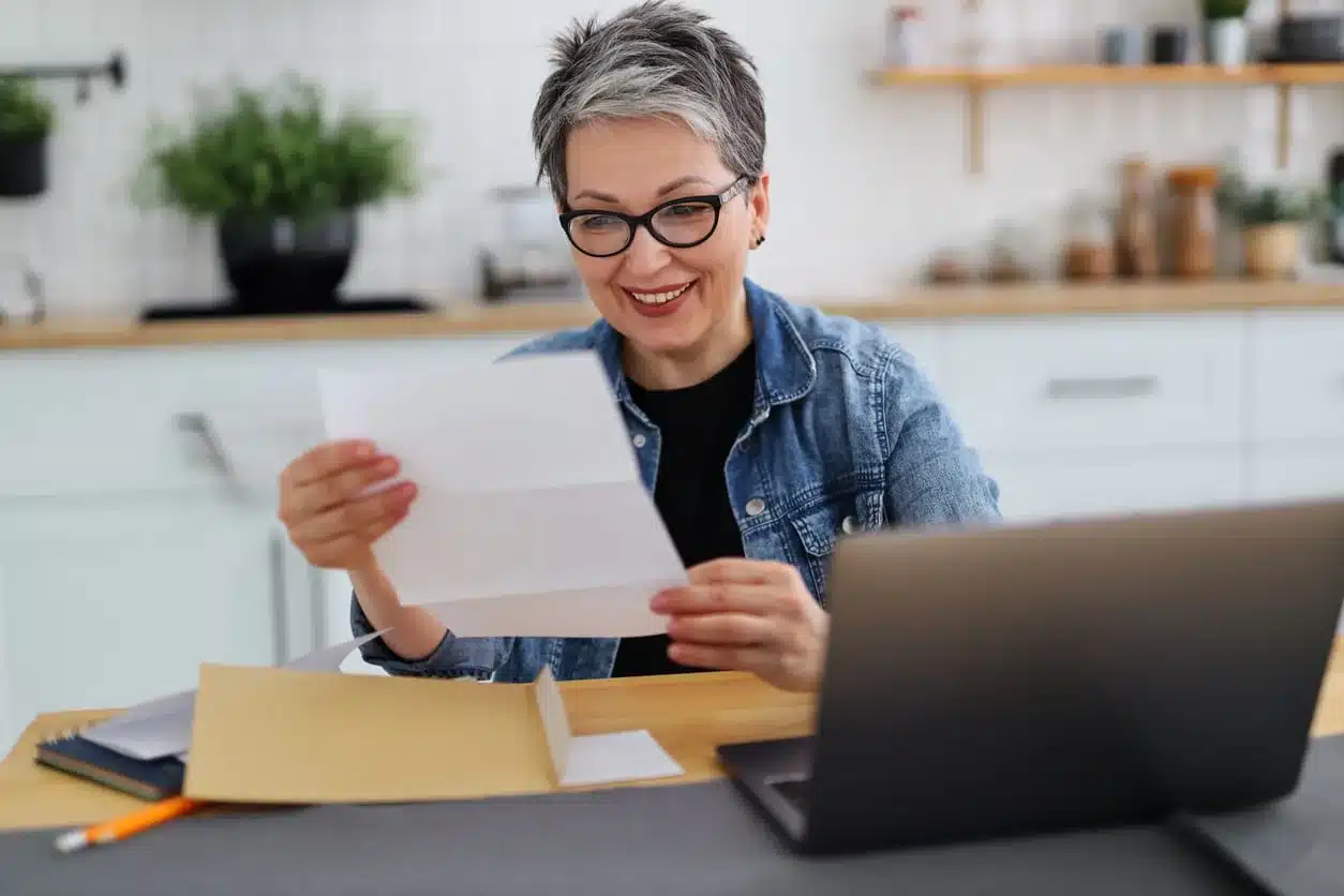 Woman with short gray hair and glasses smiling while reading a document at a home desk with a laptop and open envelope nearby.