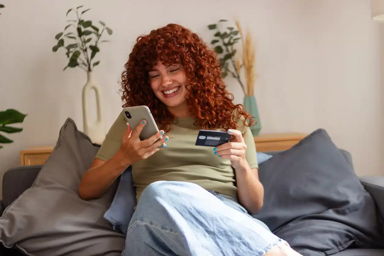 A woman relaxing on her couch, smiling while making an online payment or purchase using her phone and credit card.