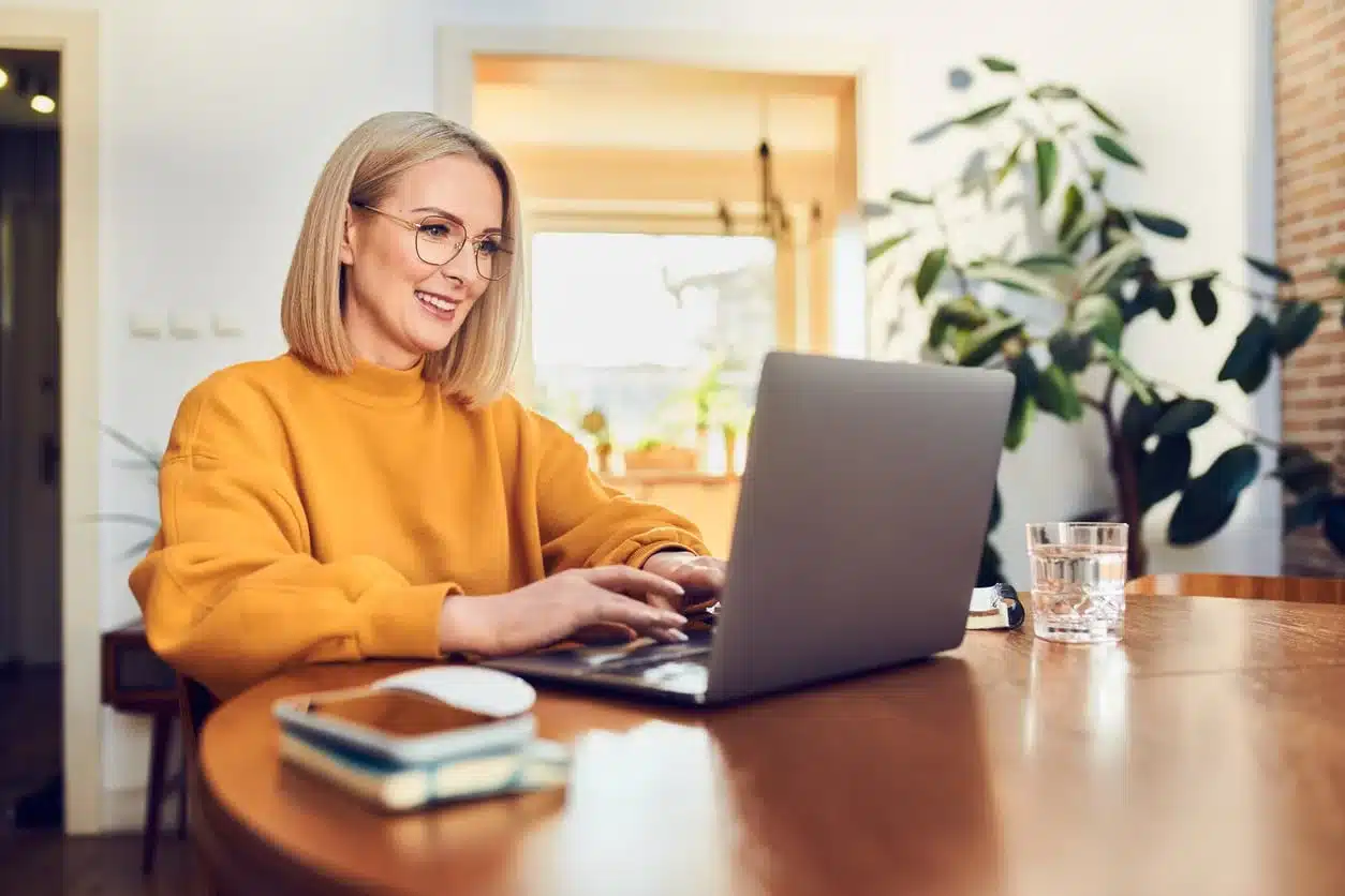 Woman in a yellow sweater smiling while working on a laptop at a wooden dining table in a bright, plant-filled home abroad.