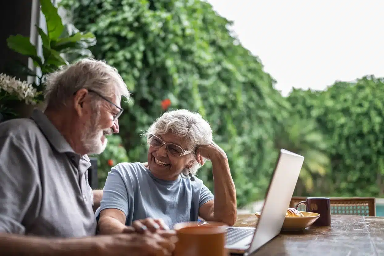Retired couple laughing together while reviewing tax information on a laptop at an outdoor patio surrounded by tropical greenery.