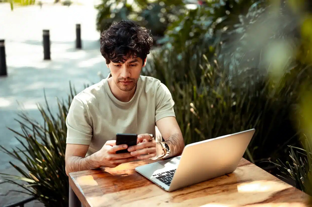 Man with curly hair checking his phone at an outdoor table with a laptop open, reviewing expat tax costs while working remotely abroad.