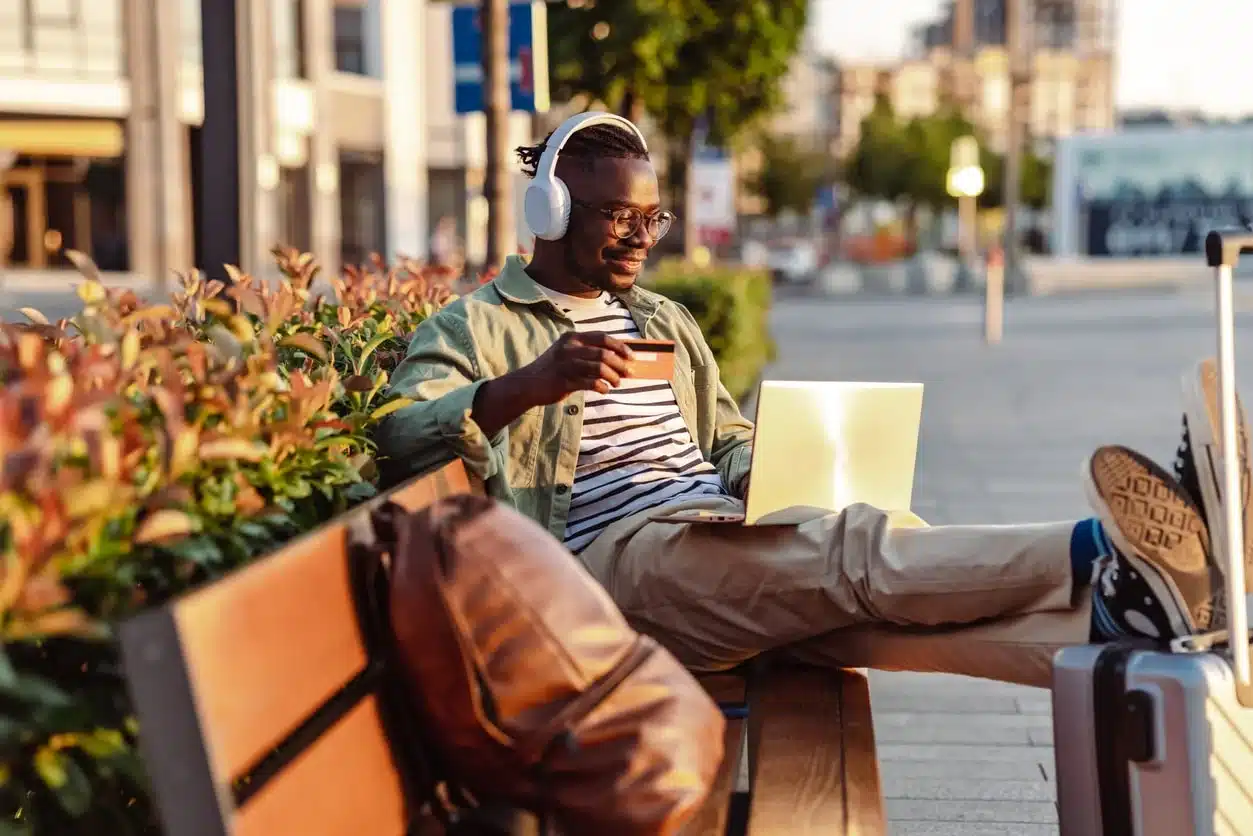 Man wearing headphones and glasses working on a laptop outdoors on a city bench, holding a card with a travel bag and suitcase beside him.