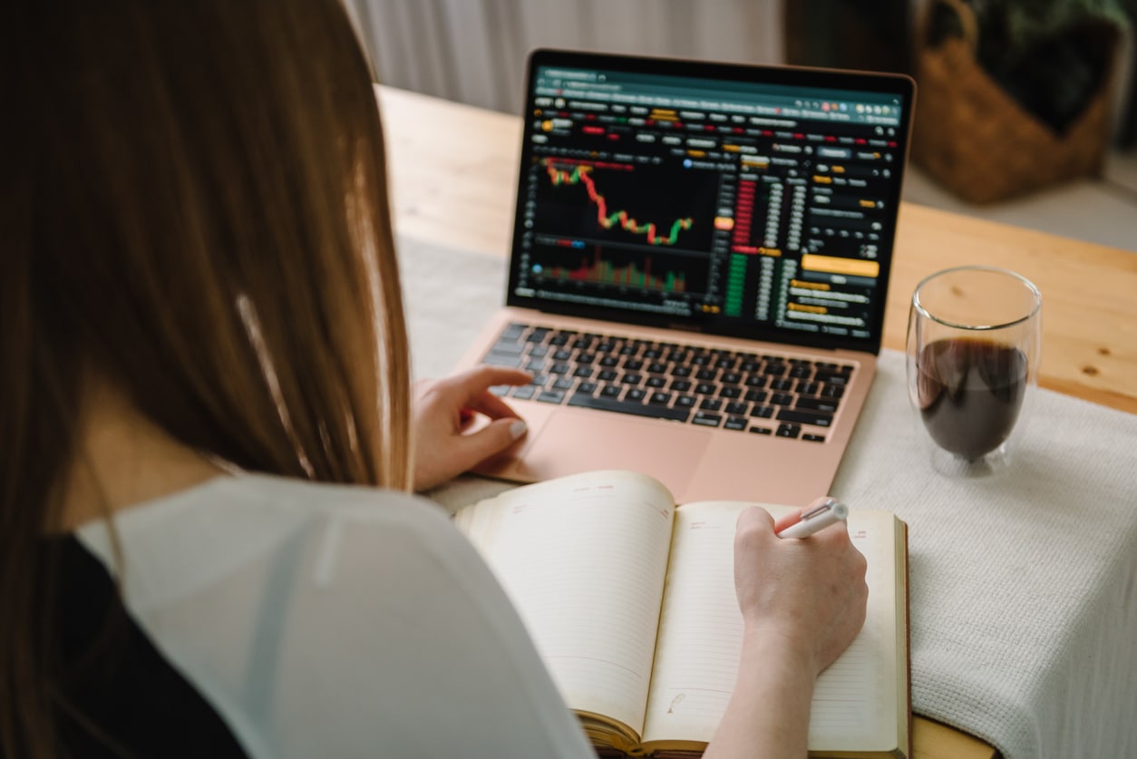 A woman reviews investment charts on her laptop while taking notes in a notebook, representing how US expats track foreign fund investments that may be classified as Passive Foreign Investment Companies for tax purposes.