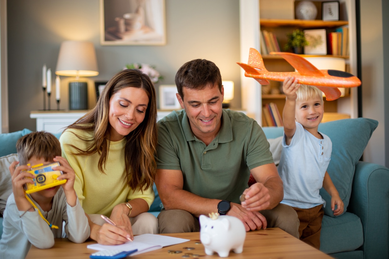 A family sits together at a table while the parents write and place coins into a piggy bank, with two young children playing nearby. The scene represents parents preparing early savings for their child, similar to opening a Trump account with the $1,000 government contribution for eligible kids.
