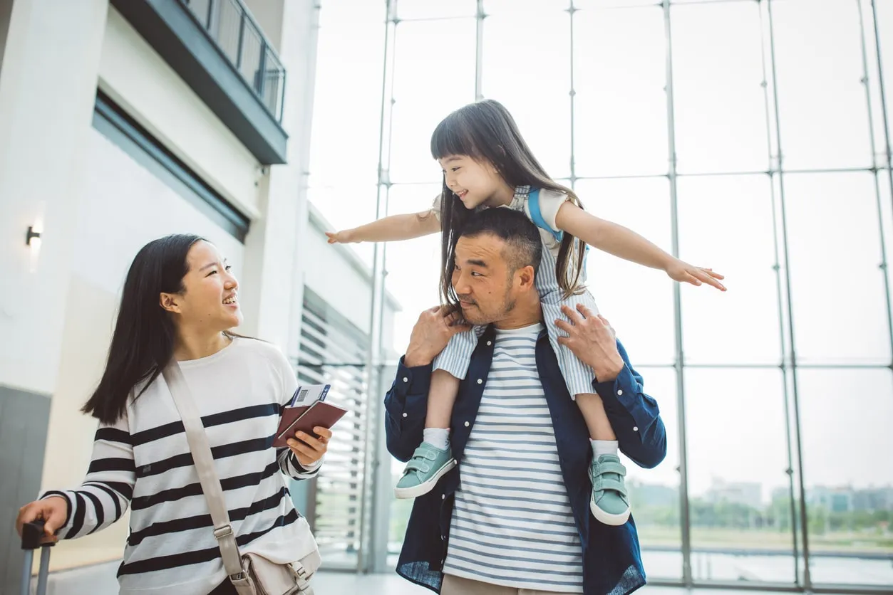 Joyful expat parent embracing adopted child at an international airport terminal with arrival screens in background.