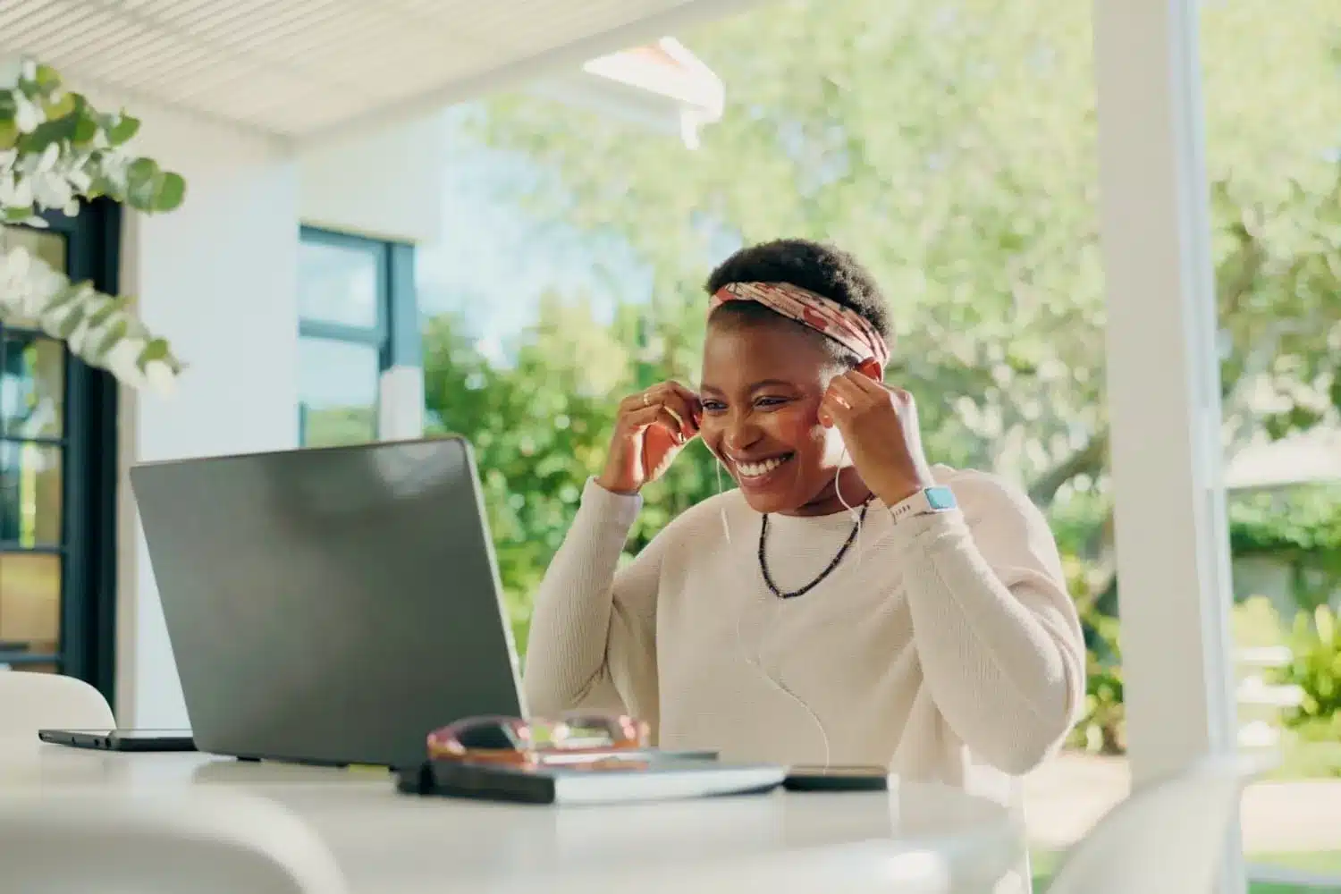 Woman on a shaded patio abroad adjusting her earbuds while working at an open laptop.