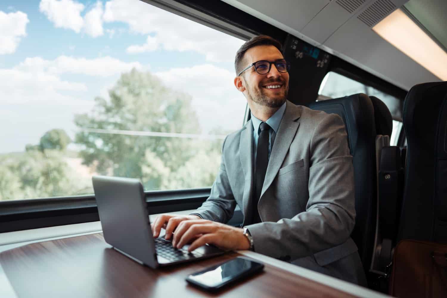 Man in a suit working at a laptop on a train, trees passing the window.