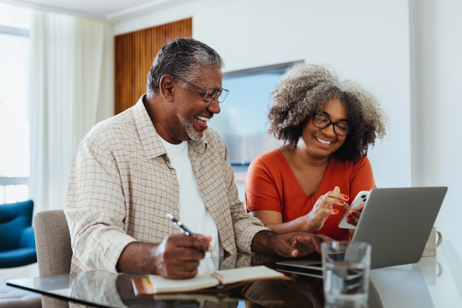 Couple at a glass table laughing over a notebook and phone, city skyline through the window.