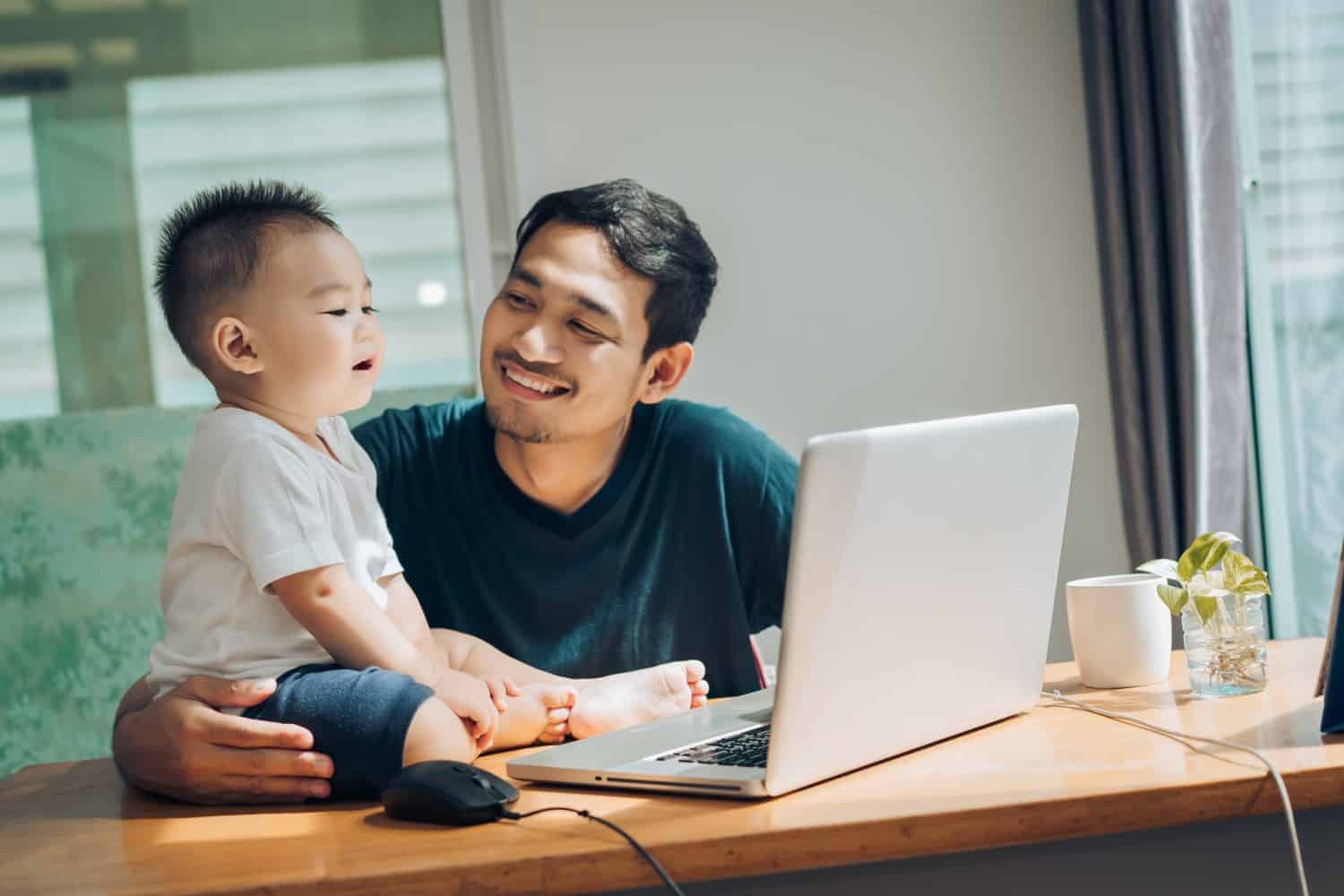 Man at a home table smiling up at his toddler beside him, an open laptop in front of them.