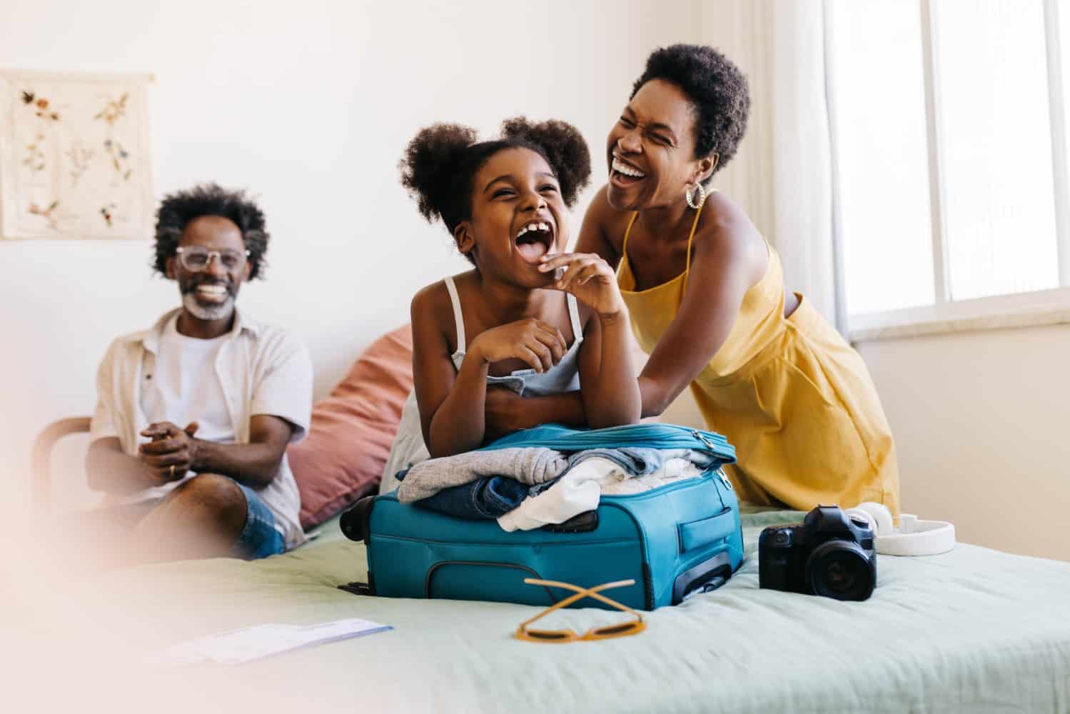 Family of three laughing on a bed around an open suitcase packed with clothes.