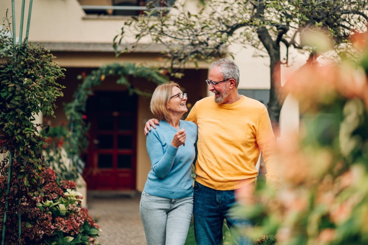 Couple walking up a garden path holding keys, a cream stucco house with a red door behind them.