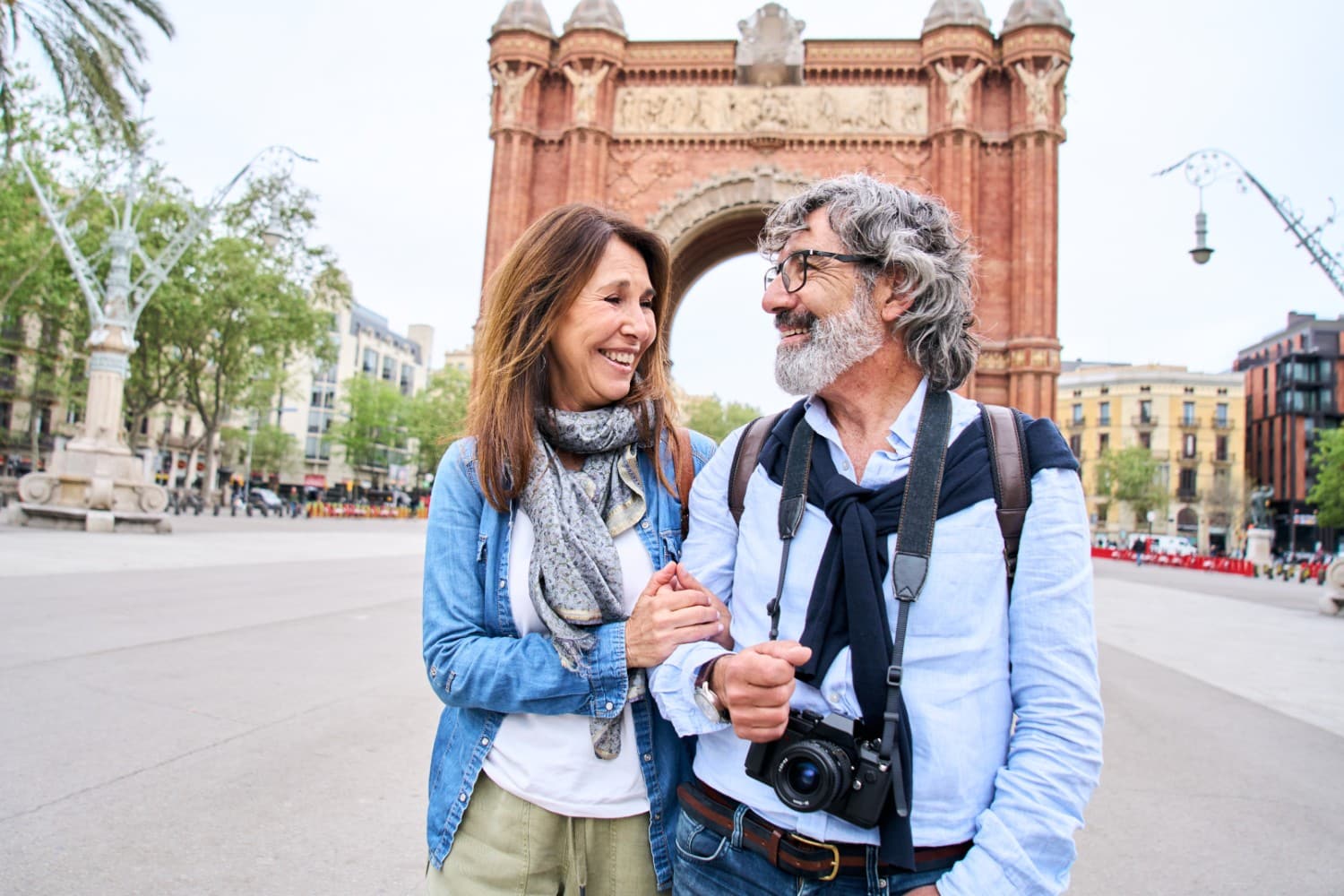 Retired couple holding hands on a plaza abroad in front of a red brick triumphal arch.
